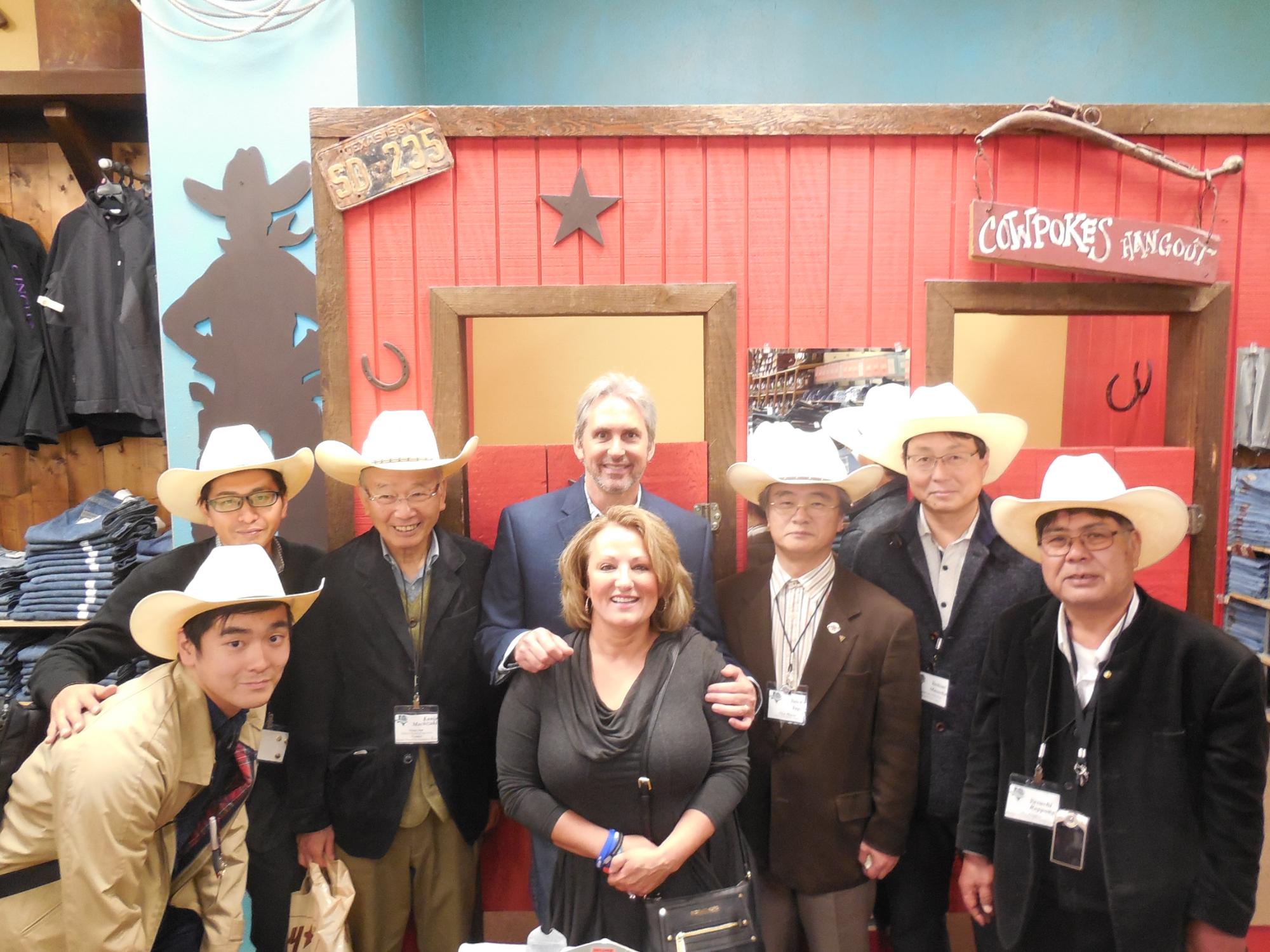 ジーンズが並べられた店舗内のカウボーイをテーマにした背景の前で、白いカウボーイハットをかぶった日本人の男性6人と、中央に立つスーツ姿の白人男性、そして手前で笑顔を見せる白人女性が集合し並んでいる写真