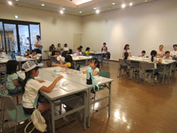 白壁の室内で、複数の子どもたちが4人ずつテーブルに座り、紙粘土で工作体験をしている様子の写真