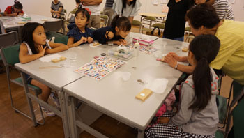 黄色のシャツを着た女性講師が熱心に指導する中、女の子たちがテーブルを囲んで座り、真剣な表情で作品を覗き込んだりしている工作風景の写真