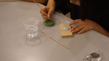 プラスチックコップに入った水と緑色の絵の具が置かれたテーブルで、女の子が割りばしの様な物を使って木片の上の白い粘土に色をつけている、創作の様子の写真