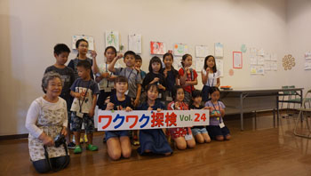 「ワクワク探検 Vol.24」と書かれた横断幕を、完成したキーホルダーを持つ大勢の子供たちが笑顔で持ってポーズをとっており、横に座るエプロンを着用した女性講師と共に記念撮影をしている写真