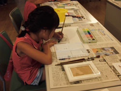 絵の具や道具が置かれた新聞紙を広げたテーブルで、ピンク色の服を着た女の子が画用紙に向かい、水彩絵の具セットと筆を使って真剣な表情で絵を描いている様子の写真