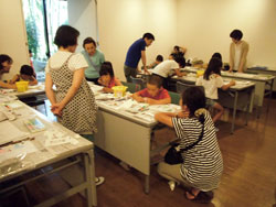 長机に座った複数の子供たちが、大人たちに見守られながら、それぞれの制作物に集中して絵を描いている様子の写真