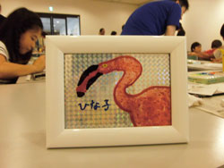 子供たちが色を塗っている机の上に、光沢のあるガラス板に、くちばしが黒く長いピンク色のフラミンゴが描かれており、「ひな子」という文字が添えられた作品が白い額縁に入れられ置かれた写真