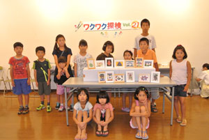 「ワクワク探検 Vol.27」と書かれた白い壁の前で、自作の額縁に入れた作品を並べた机の周りに集まった子供たちが、床に座ったり立ったりして並んでいる集合写真