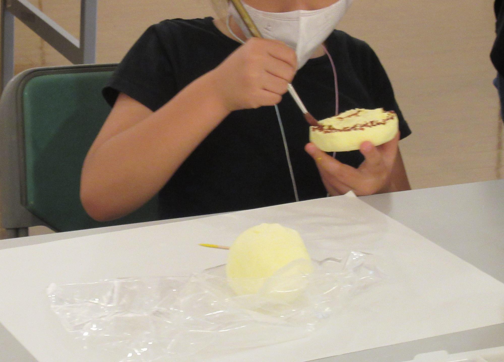 白いマスクを着け黒い服を着た子どもが、中央をくりぬいた円形の黄色い粘土に、茶色い絵の具でドーナツのようにデコレーションしている工作風景の写真