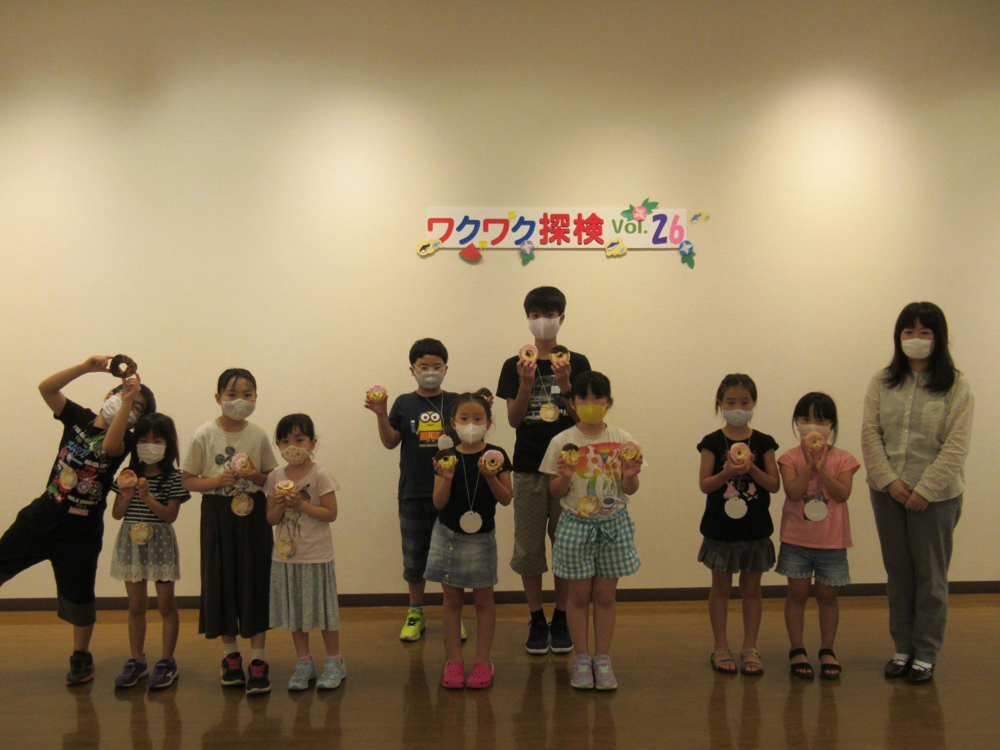 「ワクワク探検 Vol.26」と書かれた白い壁の前で、マスクを着けた白いシャツを着た女性と子供たちが、それぞれが制作したドーナツ型の作品を持って、楽しそうに記念撮影をしている集合写真