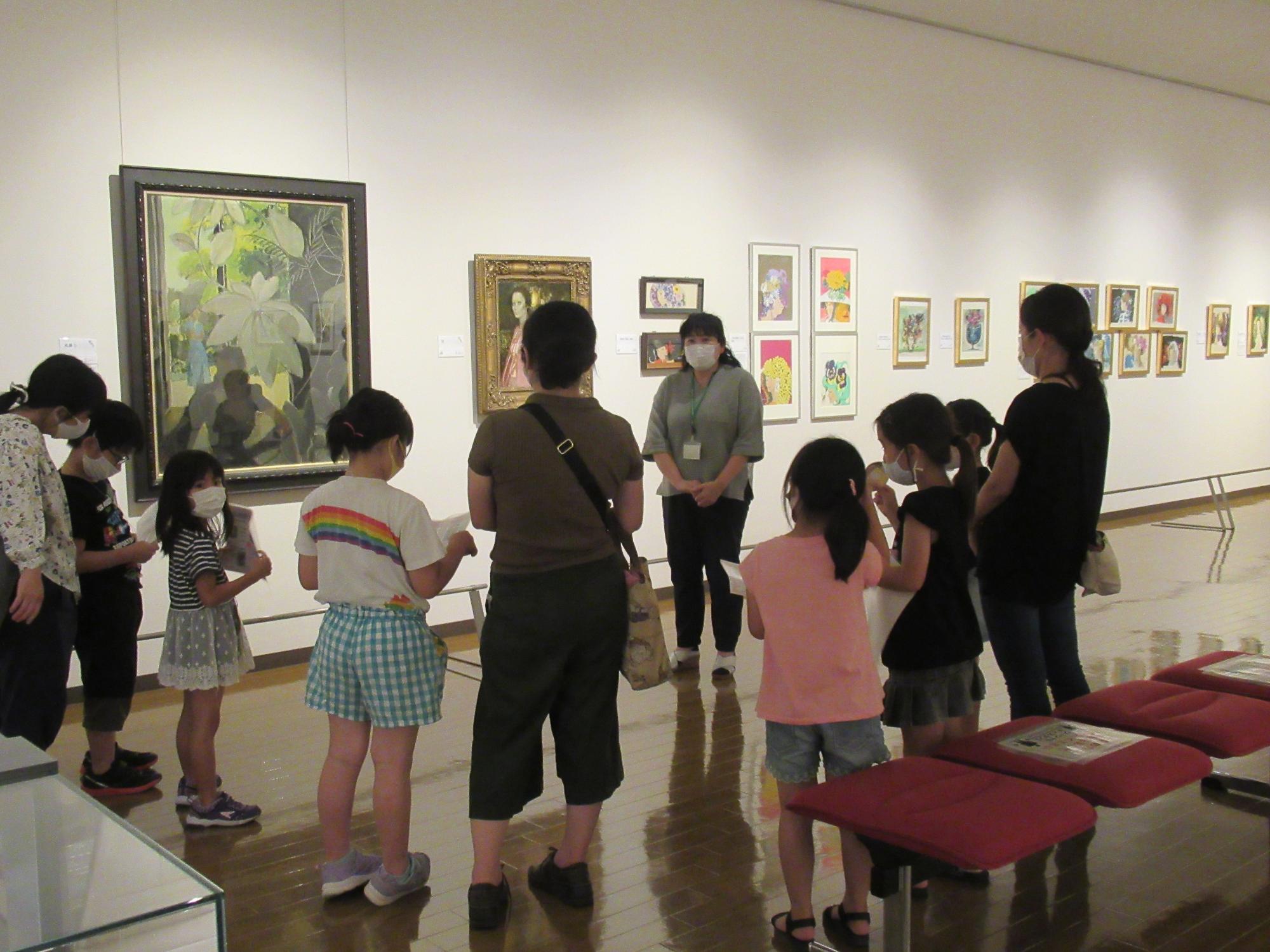 大小様々な絵画が展示された白壁の美術館の展示室で、グレー色のシャツを着た女性スタッフが、複数の親子連れの参加者に囲まれ説明している様子の写真