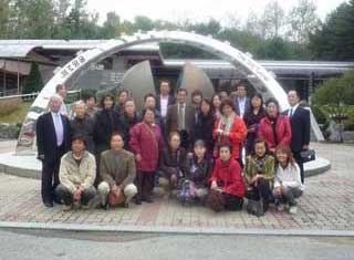 韓国の統一展望台らしき建物の前にある半円状のモニュメントの前に、数名の男性と多数の女性からなる訪問団と思われる団体が集合して記念撮影している写真