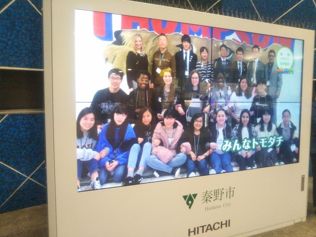 右下に「みんなトモダチ」という文字が表示された、国際交流プログラムに参加した多国籍な生徒たちと引率者たちが集合写真を撮ったものが、秦野市のロゴと日立の文字が入った大型デジタルサイネージに表示されている写真