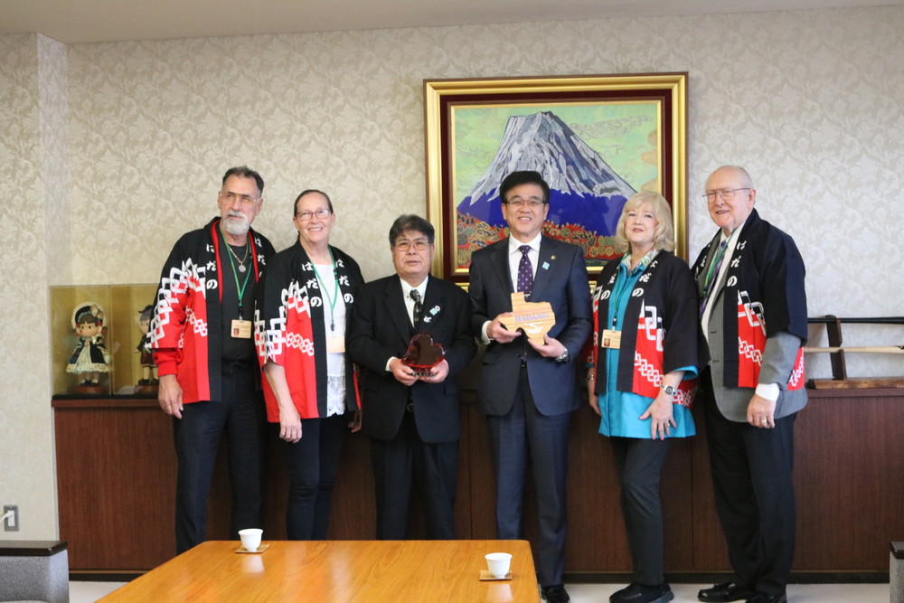 金色の額縁に入った山頂に雪が積もった大きな山の絵画が壁に飾られた落ち着いた背景の部屋で、テキサス州の形をした記念品を持ったスーツ姿の市長ともう一人の日本人男性を中央に、赤い紋が印象的な法被を着た外国人訪問団の4人が横一列に並んでいる姉妹都市交流の公式訪問の記念写真