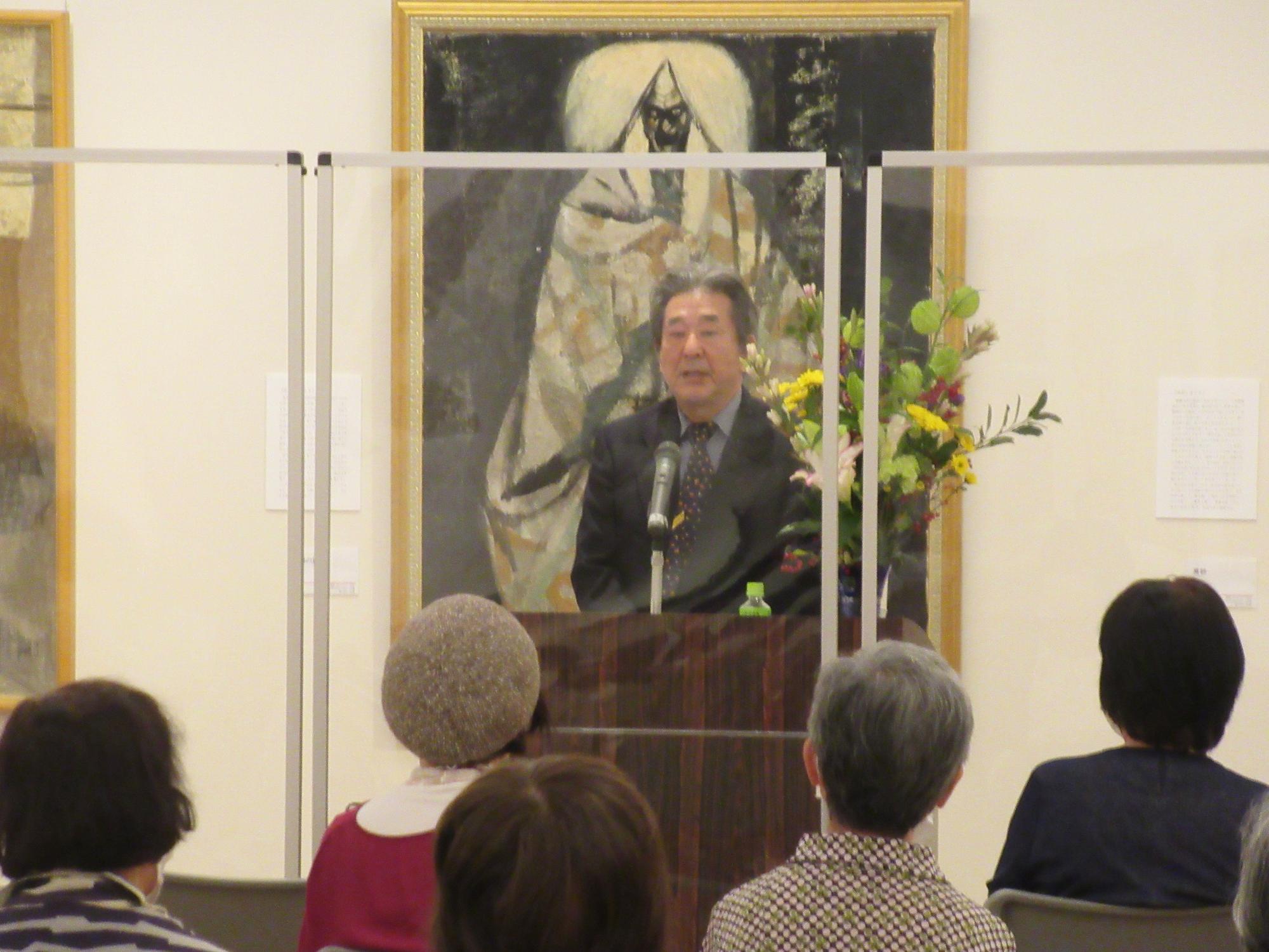 白い布のようなものを被った人物が描かれている額装された絵画を背景に、檀上に設置されたマイクスタンドの前に立つ男性が聴衆に向かって話している様子の写真