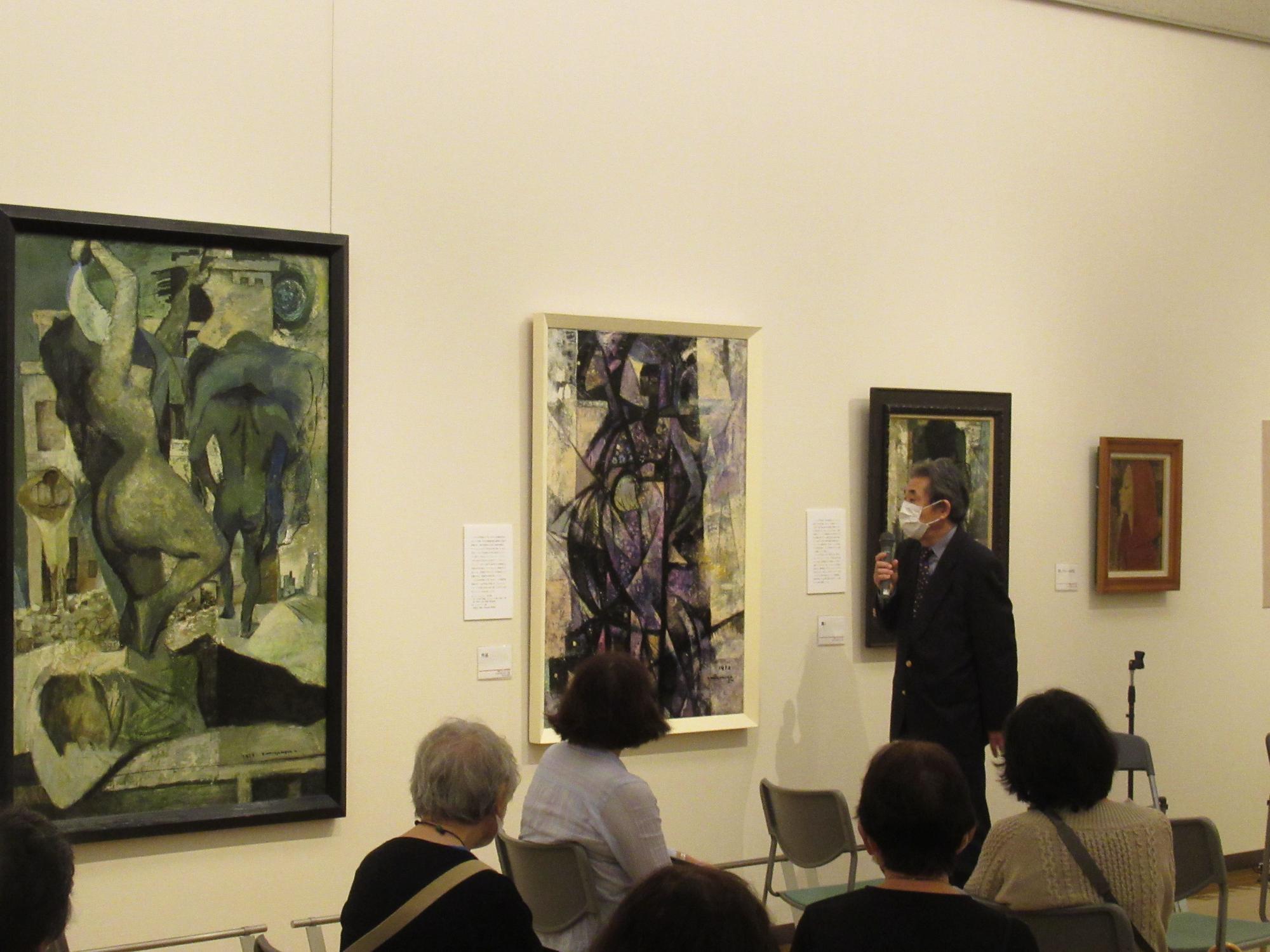 壁に大小様々な大きさの絵画が飾られた展示室で、右手でマイクを持つマスクを着用した男性が聴衆に説明している様子の写真