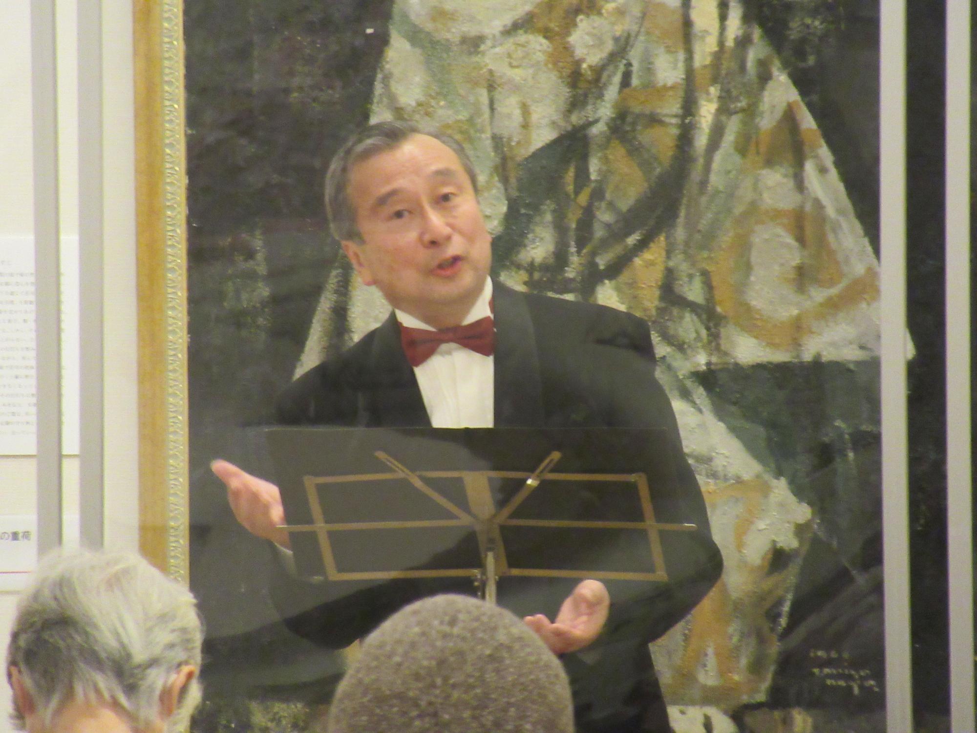 抽象的なタッチで描かれた絵画が飾られている前に立つタキシードを着て赤い蝶ネクタイをつけた男性が、譜面台を前に両手を広げて歌を披露している写真