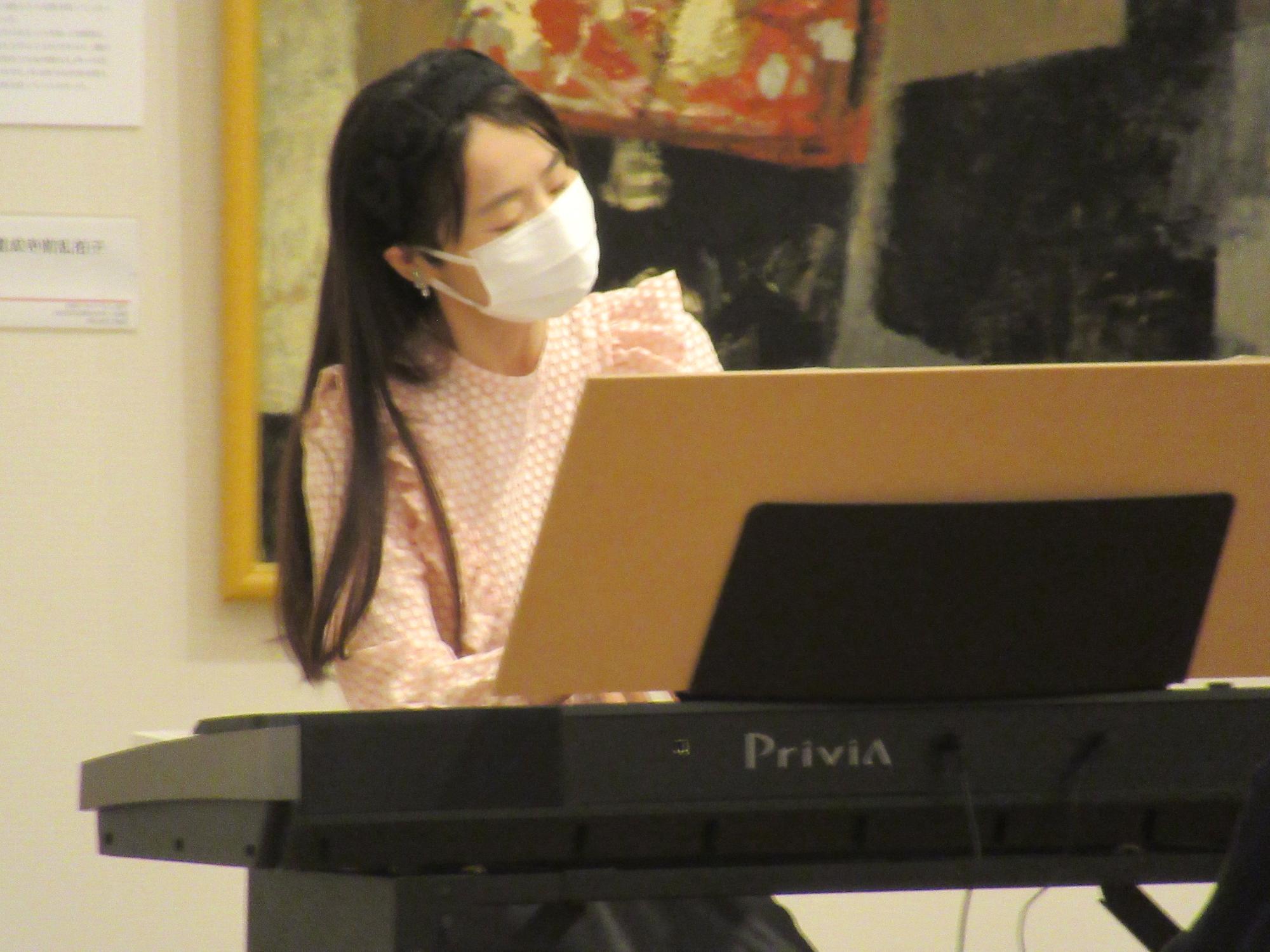 抽象的な絵画が飾られている会場で、白いマスクをつけピンク色の服を着た女性が、電子ピアノの前に座って演奏している写真