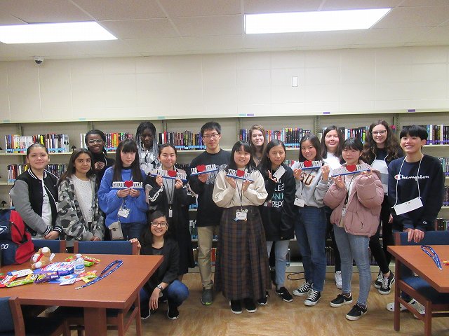 本が並んだ本棚が壁に設置された室内で、留学プログラムまたは交流イベントに参加した日本人と外国人を含む16人の若い学生たちが集合し、そのうち数人が赤い記念品を持って笑顔で立っている写真