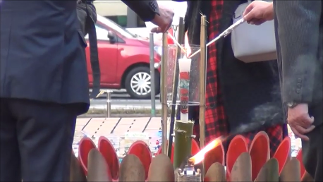 スーツ姿の2人の男性が手に持った点火棒で中央の灯篭のような装置に立てられた大きなキャンドルに火を灯そうとしている様子をクローズアップした写真（ 「市長挨拶～メインキャンドル点灯」秦野市公式YouTubeのサイトへリンク）