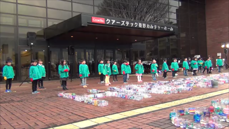 屋外で、「クアーズテック栗野カルチャーホール」と書かれた建物の入り口前に、無数の透明な容器が並べられており、お揃いの緑色の上着を着た大勢の子供たちが整列して歌を披露している様子の写真（「北幼稚園手話コーラス」秦野市公式YouTubeのサイトへリンク）