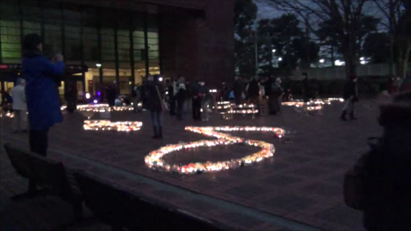 日が落ちて薄暗い広場で、地面に並べられた無数のキャンドルが明るく光り、音符の形など様々な形を曲線状に描き出しており、周囲には多くの人々が集まりその様子を見つめている写真（ 「キャンドル点灯中」秦野市公式YouTubeのサイトへリンク）