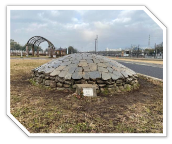 ところどころ草が生えた通路の角に「集積1」と書かれた石のプレートが置かれ、不揃いな石がドーム状に積み重ねられた屋外のオブジェの作品の写真