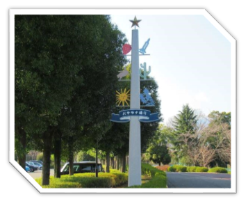 駐車場と並木道沿いの間に、上に向かって細くなっている中央部分のブルーの帯に「パサデナ通り」と書かれ、頂上に星、イチゴやサボテンなど様々な飾りがついた白い柱が立っている写真