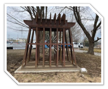 駐車場に隣接する土の広場の中央に、茶色の金属の柱で組まれた立方体の枠の中に、歯車のような金属製のオブジェが入っている作品が設置されている写真