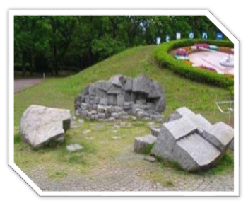 木々に囲まれた小高い広場に設置された花時計のある手前の平坦な石畳の上に、砕けた不揃いな形をした大きな岩が複数配置され、中央には積み重ねられた石ころの構造物が見える彫刻の写真