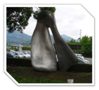 木々や生垣に囲まれた屋外に設置された、銀色に光る抽象的な形状で滑らかな曲線と光沢のある表面を持つ、2つの巨大なオブジェが上で円形になりつながっている金属製彫刻の写真