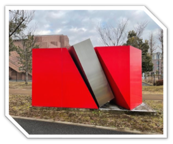 斜めに切り裂かれた赤い立方体の間に、銀色の直方体が突き刺さったような、ダイナミックな形状の現代彫刻が屋外に展示されている写真