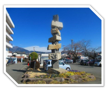 白い建物が建ち沢山の車が停まっている駐車場の敷地内に、窪みのあるいくつかの石が不安定に積み重ねられたような、高くそびえる抽象的な石の彫刻が、花壇の中心に設置されている写真