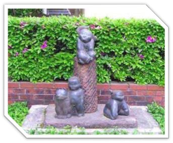 赤レンガ作りの花壇の中にピンク色の花が咲いた生垣を背景に、円柱の上に一体、その下に三体の幼い子どもや動物を思わせるブロンズ像が配置された屋外彫刻の写真