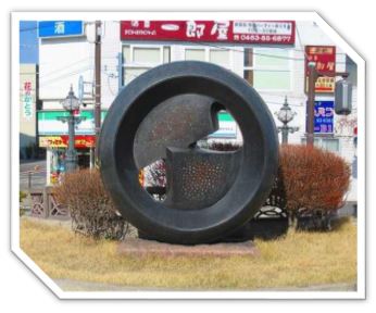 背景にコンビニが映る通りに面した小さな植え込みに、中心に穴が空いた黒いドーナツ型に、一部を切り取った金属板を組み合わせたような抽象的な現代彫刻の写真