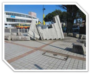 背景に駅舎が見える駅前広場の舗装された地面に並べられた、倒れそうに重なって斜めに立っている高さの違う5枚の砕けたコンクリートブロックのような抽象的な石が、L字型の石に支えられて立っている彫刻の写真