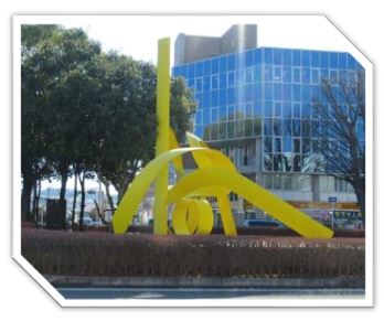 背景に全面ガラス張りのビルが立ち大きな木々や生垣に囲まれた円形の敷地の中央に、明るい黄色のリボンが絡み合ったような抽象的な現代彫刻が設置されている写真