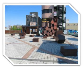 茶色い金属製の複数の直方体や立方体が、中央の巨大な半円形のオブジェから飛び出したように広場に配置された抽象的な屋外彫刻の写真