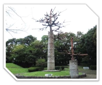 木々に囲まれた公園内に、幹は石造り、枝は鉄で出来た二本の木を模した抽象的な彫刻で、大きな彫刻が小さな石で囲まれた円形のこんもりした芝生の中央に立ち、小さな彫刻は枠の上に立ち並んでいる屋外アート作品の写真