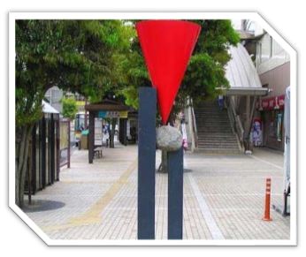 黒い二本の柱の上に、石と赤い逆さまの円錐が積み重なるように配置された、駅前広場に立つ現代彫刻の写真