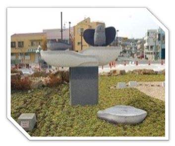 屋外の芝生広場の中心に、翼を広げた鳥と船を模した二つの石が、波打つような台座の上に配置されている彫刻の写真