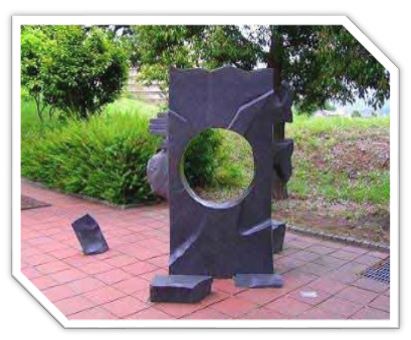 木々に囲まれた通路上に、真ん中に丸い穴が空いた黒い一枚の石板が、割れた破片とともにタイル敷きの地面に配置されている抽象的な屋外彫刻の写真