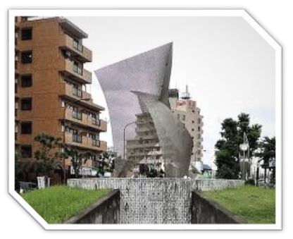 アパートやビルに囲まれた円形の広場に、背景のマンションが透けて見える薄い金属メッシュで作られた波打ちながらねじれたような抽象的なモニュメントが立っている写真