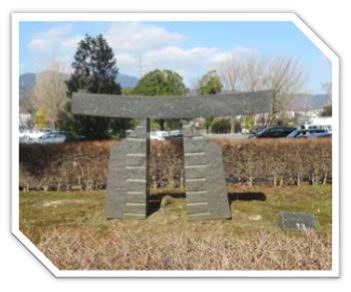 生垣に囲まれた広場に設置された、等間隔で横線が刻まれた二つの石の柱の上に、湾曲した一枚の板を載せたような鳥居を抽象化した石造りの屋外彫刻の写真