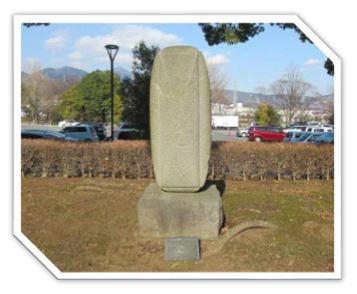 沢山の車が停まっている駐車場の傍の生垣に囲まれた広場に設置された、石の台座の上に石塔のような形をしたモニュメントが乗っている、抽象的な石造りの屋外彫刻の写真