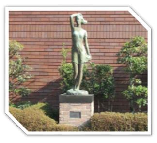 低木や生垣のあるレンガ造りの建物の前に、髪や服が風に吹かれなびいている薄い布をまとった女性が、右手を頭に置いて遠くを見つめている様子の全身像が、レンガでできた台座の上に立つブロンズ像の写真