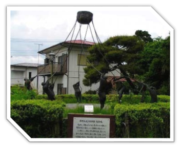 生垣に囲まれた中に設置された、重い槌を滑車で吊り上げては落とす地固めの作業を複数の人物で象ったブロンズ像の写真