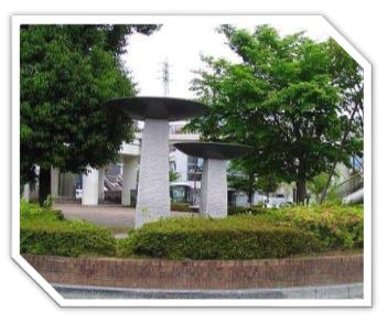 木々や生垣に囲まれた広場に設置された、異なる高さの二本の石柱の上に、黒い皿状の円盤が乗った、キノコのような形をした抽象的な屋外彫刻の写真