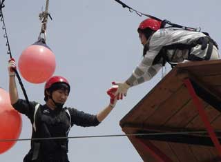 命綱やヘルメットを装着した2人の学生が高い木製プラットフォームと吊り下げられた大きなピンクのボールがある高所で手渡している様子の写真