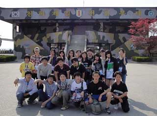 韓国語の文字が書かれた迷彩柄の建物前で、学生たちが直立したりしゃがんだりして集合写真を撮っている写真