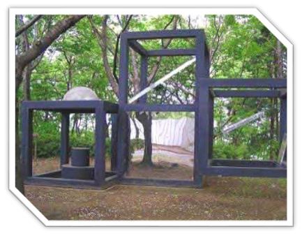 木々に囲まれた公園内に、立方体や直方体を組み合わせた黒い金属製の枠組みの中に、ドーム型の石や円柱が配置されている抽象的な屋外彫刻の写真