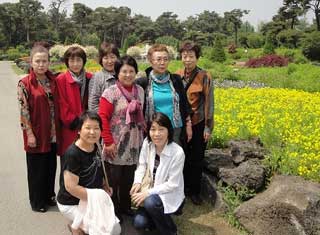 黄色い菜の花畑が広がる自然豊かな屋外の公園で、二人がしゃがみ、六人が立ってポーズをとっている、訪問団の女性たちと思われる計八名の集合写真