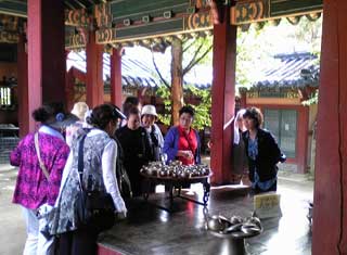 朝鮮半島の伝統的な建物の中で、数名の女性たちが中央に置かれた多数の金属製の器が載った台を取り囲んで、熱心に見学している様子の写真